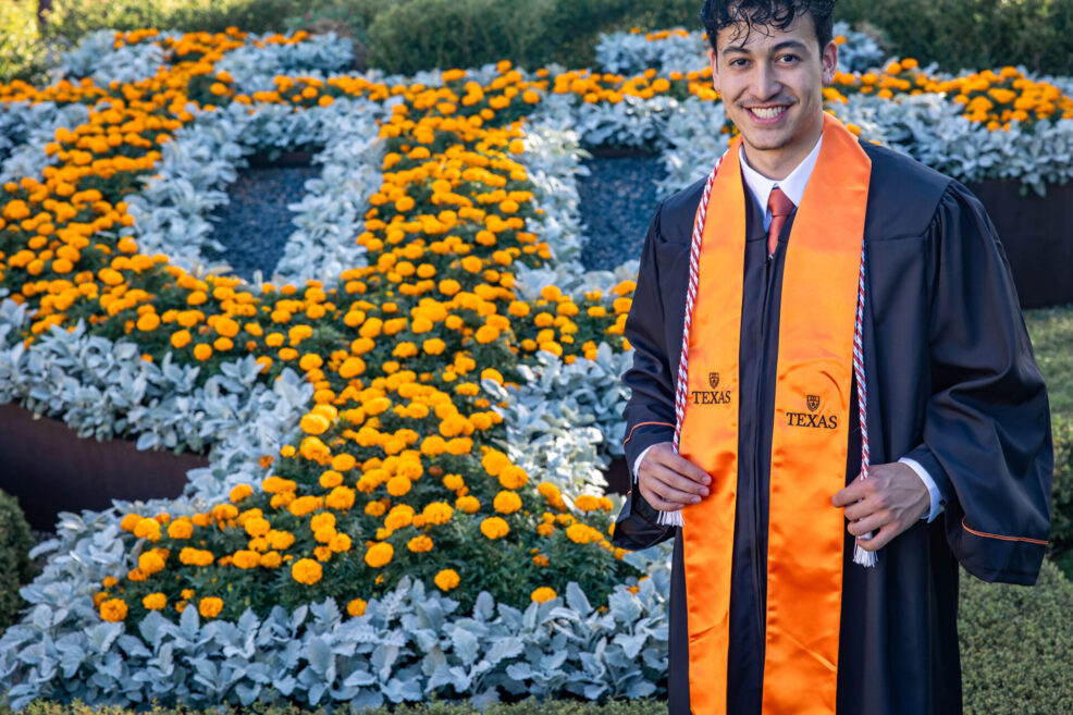 UT college senior in gown and regalia in front of flowers spelling out letters U and T